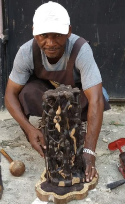 A carver kneeling beside a tall carving of intertwined figures