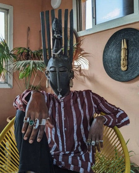 A person seated wearing a tall carved mask, a mask hung on the wall behind