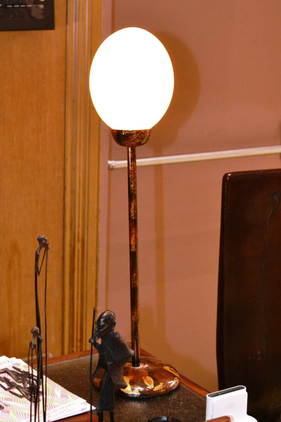 Ostrich Egg Table Lamp lit on a desk with its painted metal stem and round base beside small metal figures