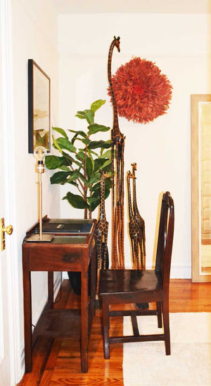 Tall and small Giraffe carvings between a wooden desk and chair, with a red feather wall hanging behind