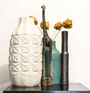 A 2 foot Giraffe on a dark side table among white, green and black vases